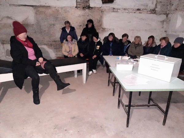 Several women sit on a bench in an underground shelter and watch as another woman demonstrates applying a tourniquet to her leg.