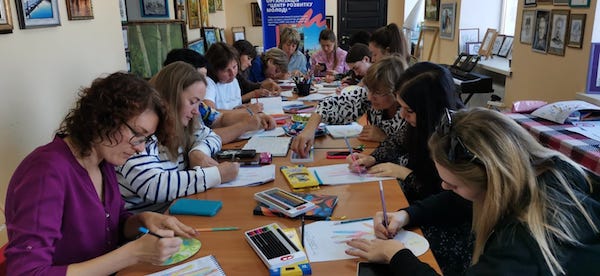 Women seated at a table focus intently as they draw on papers in an art therapy training session