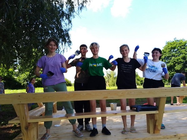 Smiling people pose with flexed arms as they paint wooden outdoor furniture