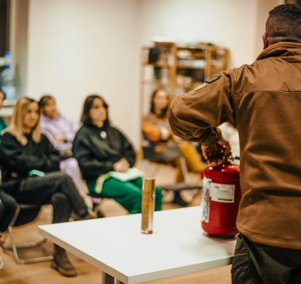 Man in brown jacket displays red fire extinguisher on table while seated audience observes