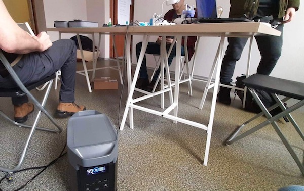 A portable power station rests on the floor next to an office table surrounded by three people. Wires connect the portable charger to devices on the table. 