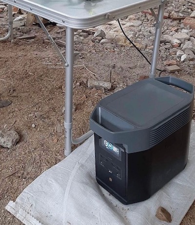 Black rectangular portable power device stands on a burlap sack on the ground next to a folding table outdoors