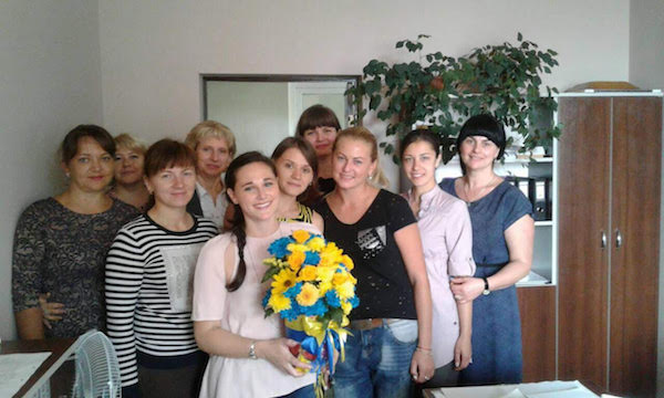 Emily (center) stands with group of women in office building. She has a bouquet of yellow and blue flowers